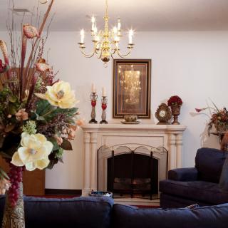 A cozy living room featuring a central fireplace adorned with decorative vases and artwork, illuminated by an elegant chandelier and complemented by floral arrangements.