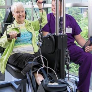 An elderly woman participates in a physical therapy session using fitness equipment, assisted by a healthcare professional.