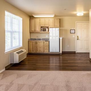 A compact apartment interior featuring a small kitchen with wooden cabinets, wood flooring, and carpeted living space.