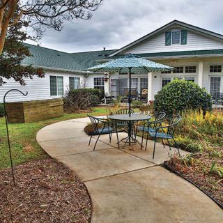 A cozy outdoor patio area with a table and umbrella, surrounded by greenery and a pathway leading to the house.