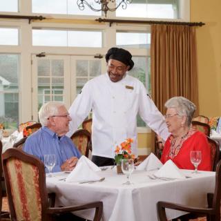 A chef interacts with a senior couple enjoying a meal in a restaurant.