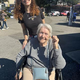 A young woman joyfully poses with an elderly lady in a wheelchair on a sunny day.