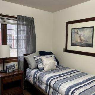 A cozy bedroom with a neatly made single bed, featuring striped bedding and decorative pillows. A nightstand with a lamp sits beside the bed, and a large painting hangs on the wall.