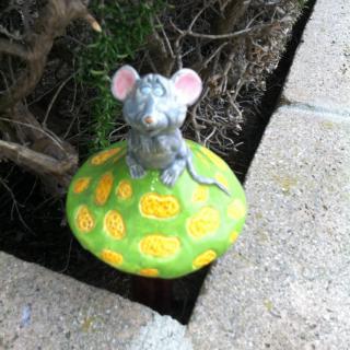 A mouse figurine sitting on a colorful mushroom in a garden setting.