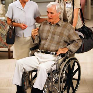 An elderly man in a wheelchair being assisted by healthcare workers in a hospital setting.