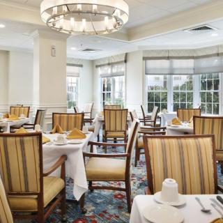 A well-lit dining room with neatly arranged tables and chairs, featuring large windows and a decorative chandelier.