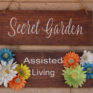 A wooden sign decorated with colorful flowers, displaying 'Secret Garden Assisted Living'.