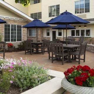 An outdoor patio area with tables and chairs under blue umbrellas, surrounded by flowering plants and adjacent to a building.