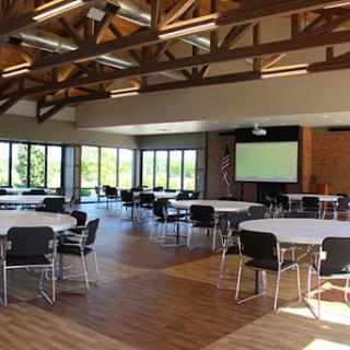 A spacious event space featuring round tables and chairs, large windows that provide natural light, and a projector screen for presentations.