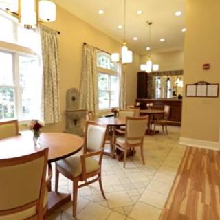 A well-lit dining area with several round tables and chairs, large windows with curtains, and warm lighting.