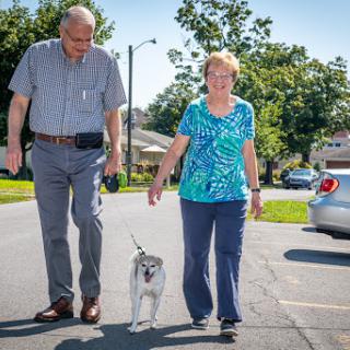 An elderly couple enjoys a sunny walk in their neighborhood with their dog.