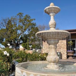 A stone fountain in a garden setting, surrounded by greenery and flowers, under a clear blue sky.
