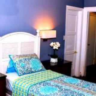 A cozy bedroom with blue walls, featuring a single bed adorned with colorful floral bedding and a nightstand with a vase of flowers.