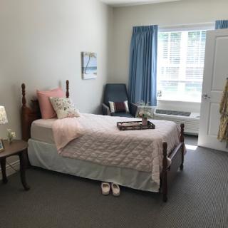 A cozy and well-lit bedroom featuring a single bed with soft pink bedding, a nightstand, and a comfortable chair.