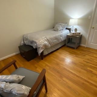 A minimalist bedroom with a single bed, nightstand, and chair on hardwood flooring.