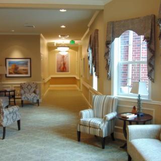 A warmly lit hallway featuring comfortable chairs, large windows with drapes, and decorative paintings on the walls.