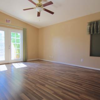 A spacious empty room with hardwood flooring and a ceiling fan, featuring large windows allowing natural light.