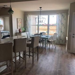 A bright and airy dining area connected to the kitchen, featuring a large window with curtains, a dining table with chairs, and a bar counter with stools on a hardwood floor.
