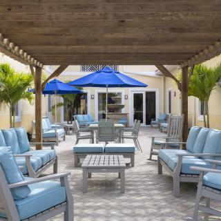 A cozy outdoor patio with cushioned seating under a wooden pergola and blue umbrellas, surrounded by palm trees.