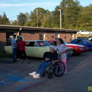 A group of people at an outdoor car show with classic cars and a person in a wheelchair.