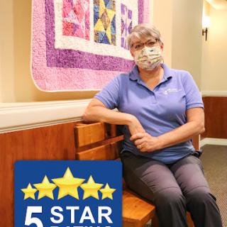 A masked individual seated in a welcoming indoor space, featuring colorful quilts and a prominent 5-star rating.