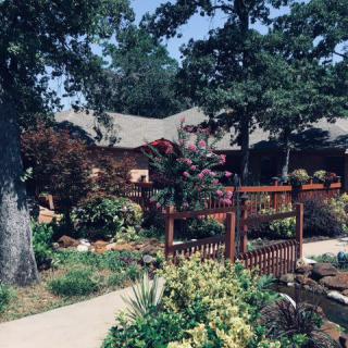 A picturesque garden with a wooden fence and a house in the background, surrounded by trees and lush plants.