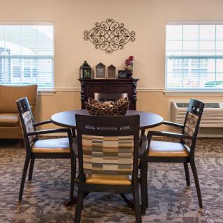 A cozy dining area with a round table and four chairs, decorated with wall art and lit by natural light from two windows.