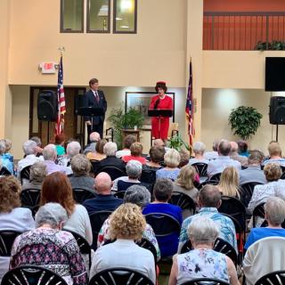 Two speakers addressing an audience seated indoors with flags in the background.