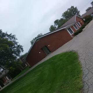 A peaceful residential area featuring a brick house surrounded by green grass and trees under a cloudy sky.