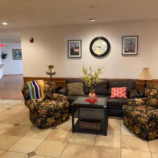 A cozy lobby seating area with a sofa, armchairs, and a table adorned with flowers, under a wall clock and framed pictures.
