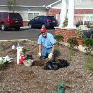 A man is gardening outside a building, surrounded by plants and tools.