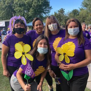 A cheerful group of participants in purple shirts holding flower props, gathered at an outdoor event.