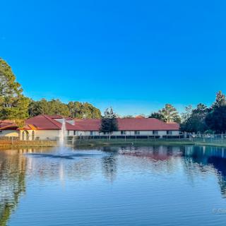 A scenic view of a serene lake with a fountain surrounded by lush greenery and charming buildings under a clear blue sky.