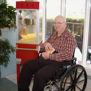 An elderly man enjoys popcorn while seated in a wheelchair next to a popcorn machine.