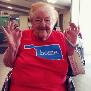 An elderly woman in a red shirt with the word 'home' and an outline of Oklahoma, smiling and giving a thumbs-up from her wheelchair.
