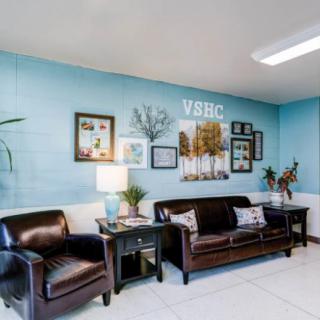 A cozy waiting room with a blue wall, featuring a sofa, armchairs, plants, and decorative wall art.