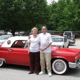 A couple stands next to a red vintage convertible in a parking lot.