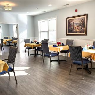 A bright dining area with neatly arranged tables and chairs, featuring a large painting on the wall and ample natural light from the windows.