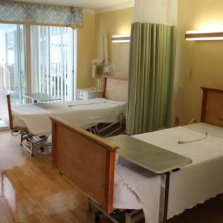 A view of a hospital patient room with two beds, curtains for privacy, and natural light coming in from the window.