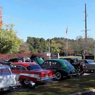 A gathering of classic cars showcased under a clear blue sky, with an American flag waving in the background.