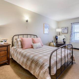 A cozy and well-lit bedroom featuring a metal-framed bed with plaid bedding and pink accent pillows, flanked by a wooden nightstand and lamp. A comfortable chair sits by the window, adding charm to the room's decor.