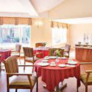A cozy dining room with tables set for a meal, featuring red tablecloths and ample natural light.
