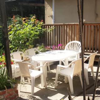 A sunny outdoor patio featuring a white plastic table and chairs surrounded by greenery and flowers.