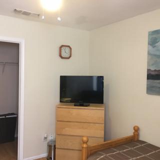 A bedroom with a television on a dresser, a clock on the wall, and a bed in the corner.