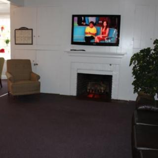 A cozy living room with a television mounted above a fireplace, featuring comfortable chairs and indoor plants.