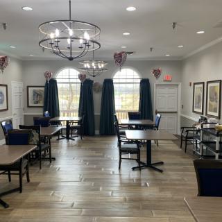 A well-lit dining room with neatly arranged tables and chairs, elegant chandeliers, and large windows adorned with curtains.