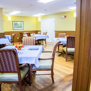 A well-lit dining room with neatly arranged tables and chairs, featuring tablecloths and a warm atmosphere.
