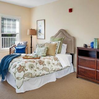 A cozy bedroom with a neatly made bed, decorative pillows, a dresser, and a window letting in natural light.