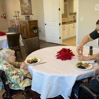 An elderly person and a caregiver sharing a meal at a dining table in an indoor setting.