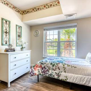 A bright and cozy bedroom with floral bedding, featuring a white dresser and large window allowing natural light to flood the space.